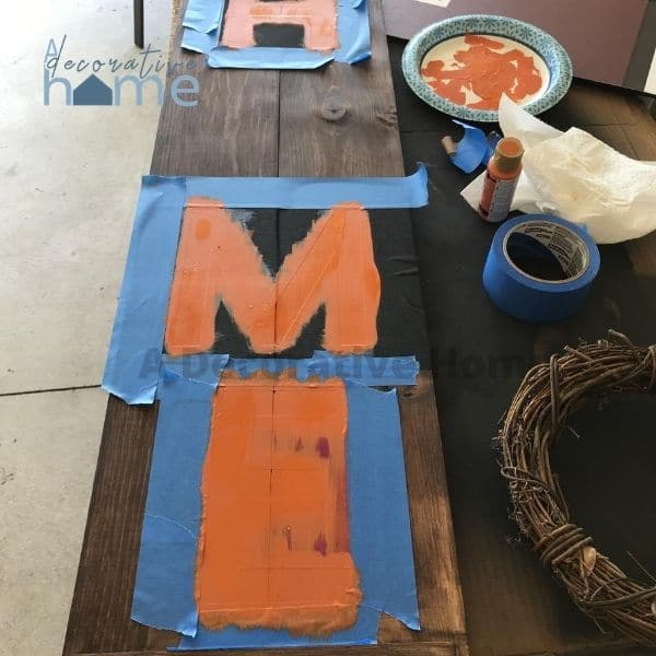 The wood board that has the letters painted on it with blue painters tape on it.
